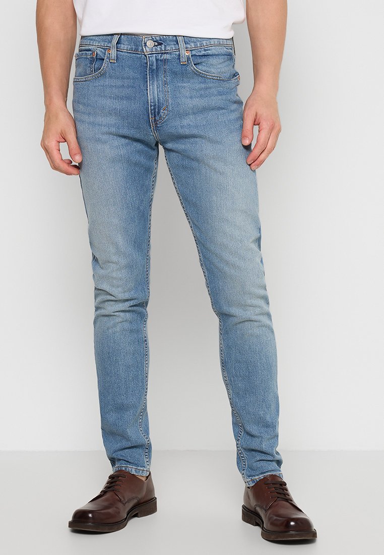 Jeans en denim bleu clair avec une coupe slim, cinq poches, des coutures contrastantes et une fermeture à bouton classique. Portés avec des chaussures marron foncé.