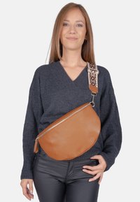 Sac bandoulière en cuir marron de forme arrondie, fermeture éclair argentée et bandoulière à motif. Le sac présente une surface texturée et un détail de frange.