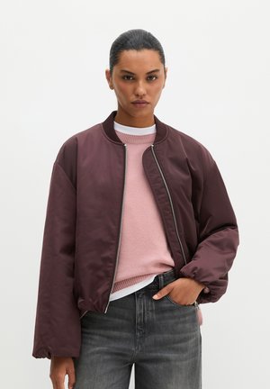 Woman with slicked-back hair wears a maroon bomber jacket over a pink sweater and black jeans, standing with one hand in her pocket.