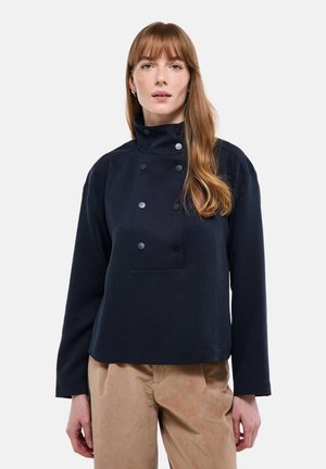Femme aux cheveux longs châtain clair portant une veste bleu marine à col montant boutonnée et un pantalon beige, debout devant un fond blanc uni.