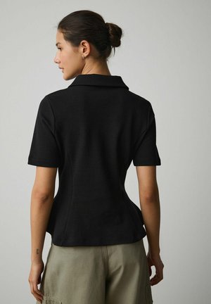 Moteris su tamsiais plaukais surištais į kuodą, vilkinti juodą trumpomis rankovėmis marškinių tipo palaidinę ir šviesiai chaki spalvos kelnes, stovinti nugara priešais paprastą pilką foną.