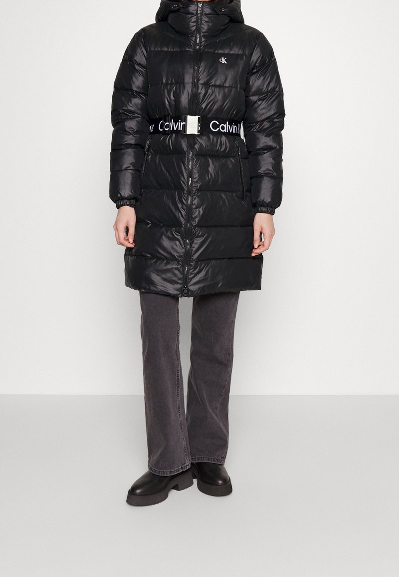 Czarna puchowa kurtka z kapturem, pikowana, z metalowym paskiem z logo. Nosić na szare spodnie i czarne buty.