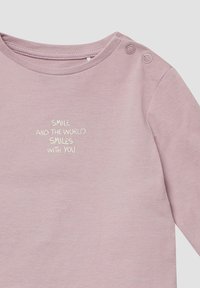 Roze katoenen top met lange mouwen en ronde hals, met twee drukknopen op de schouders en geborduurde tekst "LAAT EEN GLIMLACH ZIEN EN DE WERELD GLIMLACHT MET JE MEE."