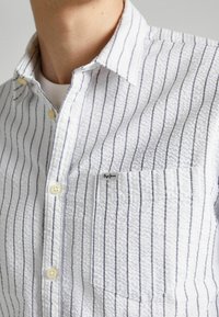 Camisa blanca de botonadura con textura y rayas verticales azules, que incluye un bolsillo en el pecho y una pequeña etiqueta de la marca cerca del bolsillo.