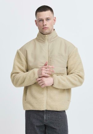 Giacca beige con maniche in pile morbido e testurizzato e un corpo liscio. Presenta un colletto alto e una tasca frontale. Chiusura con zip.