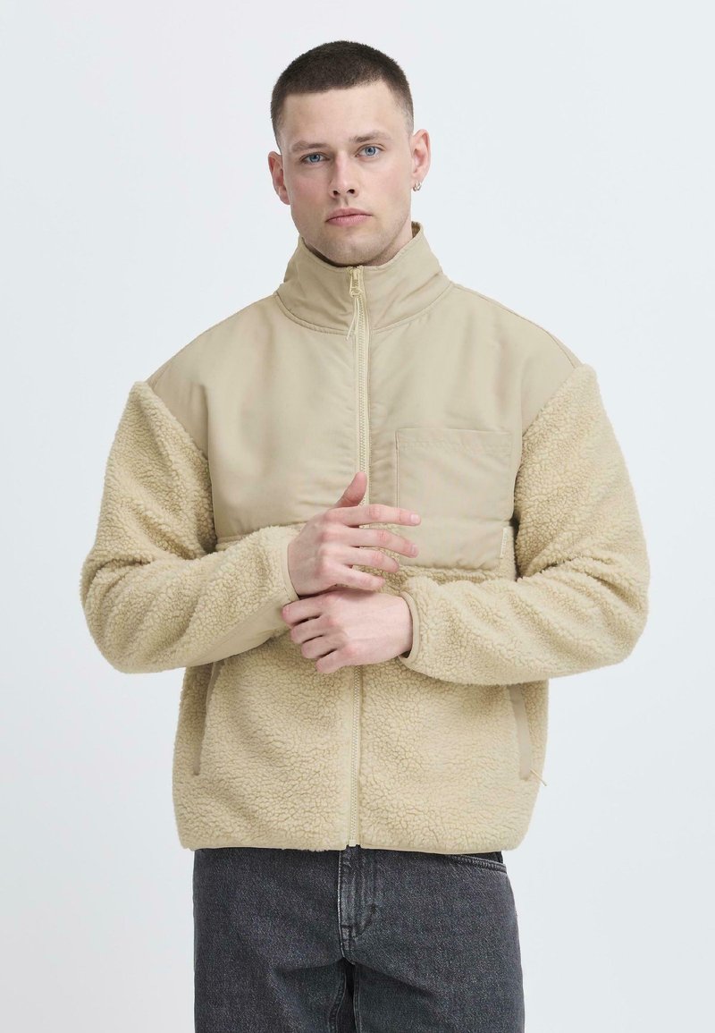 Giacca beige con maniche in pile morbido e testurizzato e un corpo liscio. Presenta un colletto alto e una tasca frontale. Chiusura con zip.