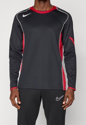 Person trägt ein schwarz-rotes Sportshirt mit langen Ärmeln und schwarze Hose mit weißen Nike-Logos, steht vor einem einfarbigen Hintergrund.