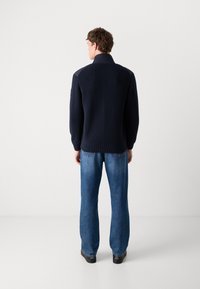 Marineblå strikkegenser med høy krage og strukturert mønster, kombinert med lyseblå rette jeans og svarte sko, sett bakfra.