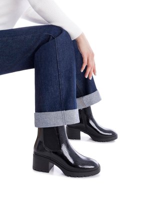 Botas de tobillo negras y brillantes con un tacón grueso, paneles laterales elásticos y suela texturizada, que se llevan con jeans azules recortados con puños doblados.