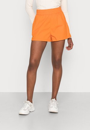 Pantalones cortos deportivos naranjas con cintura elástica, confeccionados en un material ligero, combinados con zapatillas gris claro sobre un fondo gris claro.