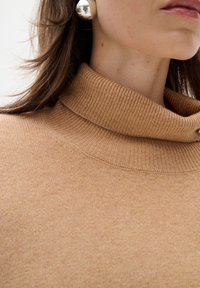 Pulover bej cu guler tip turtle, realizat dintr-un material moale. Textura netedă notabilă și detaliile cusăturii vizibile aproape de decolteu.
