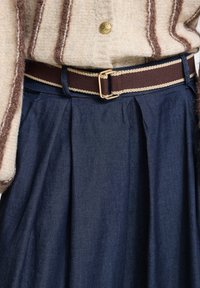 Κρεμ πλεκτό πουλόβερ με καφέ ρίγες, μπαίνει μέσα σε σκούρα μπλε πλισέ φούστα στερεωμένη με καφέ ζώνη με μπεζ άκρα και χρυσή αγκράφα.