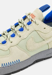 Gros plan d'une basket Nike beige et bleue montrant un tissu texturé, des panneaux en suède, des lacets à motifs, des passants de lacets bleus et une semelle à carreaux grise.