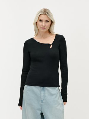 Frau mit blondem Haar, die ein schwarzes Langarmshirt mit kleinem Schlüsselloch-Ausschnitt und hellblaue High-Waist-Jeans trägt, stehend vor weißem Hintergrund.