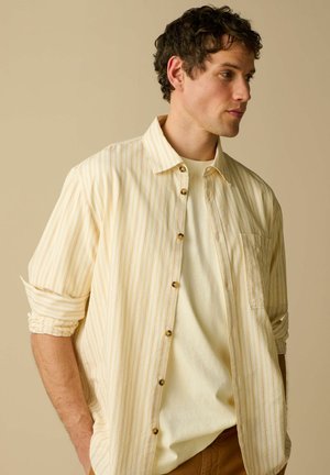 Man met krullend donker haar, gekleed in een beige gestreept overhemd met knopen over een crèmekleurig T-shirt, met opgerolde mouwen en handen in de zakken.