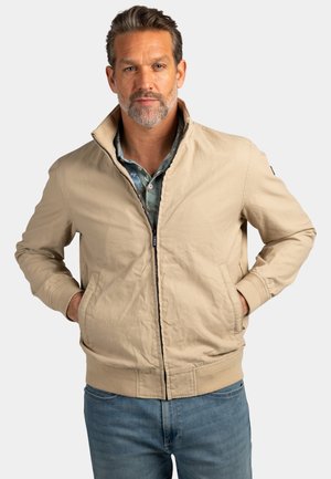 Man met een zilvergrijze baard, gekleed in een beige jas met rits over een blauw overhemd met patroon en een spijkerbroek, handen in de jaszakken, neutrale achtergrond.