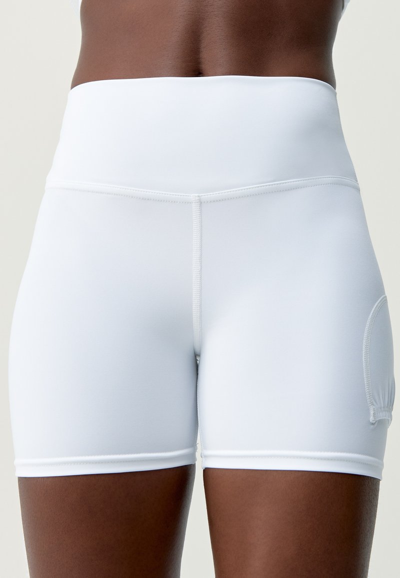 Shorts de sport blancs taille haute en tissu extensible. Ils présentent une coupe ajustée, des coutures plates et une petite poche latérale.
