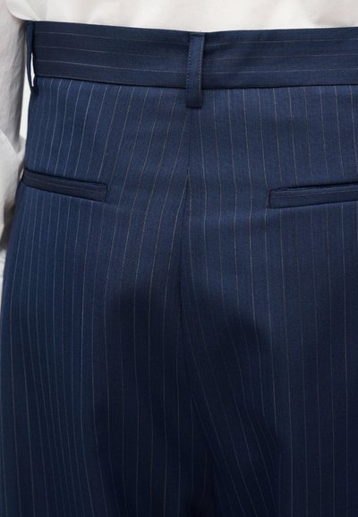 Pantaloni bleumarin cu dungi fine, buzunare cu tiv pe partea din spate și bucle pentru curea, purtați de o persoană cu un top alb.