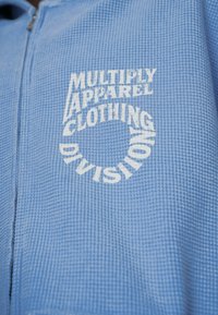 Giacca zip-up azzurro chiaro realizzata in tessuto testurizzato, con testo stampato in bianco: "MULTIPLY APPAREL CLOTHING DIVISION" disposto in forma circolare.
