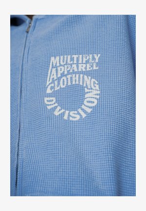 Giacca zip-up azzurro chiaro realizzata in tessuto testurizzato, con testo stampato in bianco: "MULTIPLY APPAREL CLOTHING DIVISION" disposto in forma circolare.