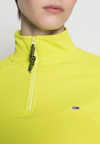 Yellow ribbed quarter-zip pullover with black zipper detail and black drawstring. Features small fabric logo on the chest.