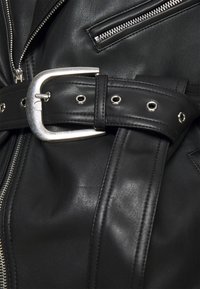 Giacca di pelle nera con cerniera argentata, ampia cintura con grandi occhielli e grande fibbia metallica. Tessuto liscio con cuciture dettagliate.