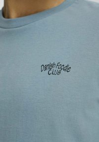 Lyseblå bomulds t-shirt med korte ærmer og rund hals, med sort broderet tekst "Danish Foodie Club" på fronten.