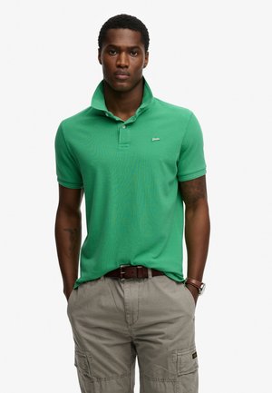 Grøn polo med korte ærmer, en spids krave og en diskret logo. Båret med lyse beige cargoshorts og et brunt bælte.