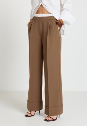 Donna che indossa pantaloni marroni a gamba larga con orli risvoltati, sandali neri con tacco, camicia bianca corta e braccialetti di legno ai polsi.