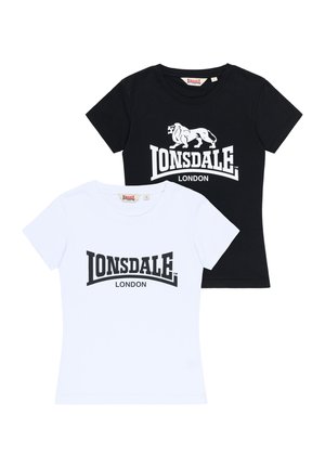Vrouw draagt een zwart Lonsdale London T-shirt, blauwe wijde jeans en witte sneakers, staat met een lichte neiging naar één kant.