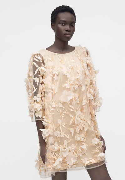 Γυναίκα που φοράει ένα διάφανο μπεζ φόρεμα διακοσμημένο με 3D λουλουδένια εφαρμογές και κεντήματα, στέκεται μπροστά σε απλό λευκό φόντο.