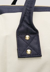 Granatowy uchwyt z ozdobnymi złotymi napami, przyszyty do teksturowanego kremowego tła, z geometrycznym wzorem ściegów.