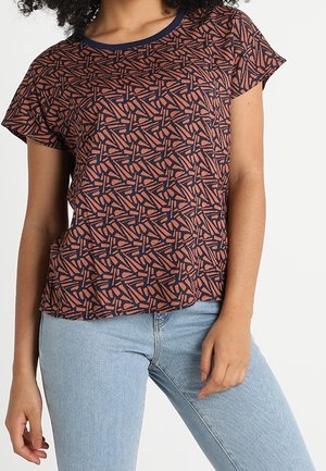 Kvinde iført kortærmet skjorte med navy- og rustfarvet geometrisk mønster og lyseblå jeans, stående mod en ensfarvet hvid baggrund.