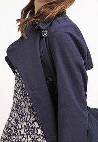 Námořnický modrý trench coat s širokými klopami, knoflíkovými uzávěry a texturovanou látkou. Nositý přes šaty s krajkovým vzorem v kontrastních barvách.
