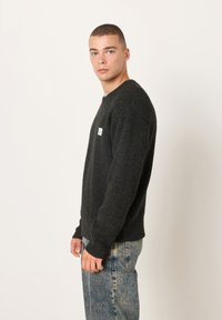 Tamnosivi rebrasti džemper s okruglim izrezom i dugim rukavima, s malim zelenim logotipom na lijevoj strani prsa. Nošen uz svijetle traperice.