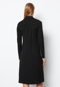 Kvinna med rakt ljust brunt hår bär en långärmad svart klänning som går ner till knäna, stående vänd mot en vit vägg.