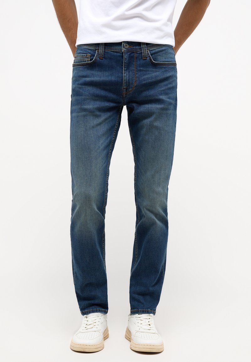 Kék farmernadrág slim fit szabással, a combján fakuló részletekkel, öt zsebes dizájnnal és narancssárga varrással. Fehér tornacipővel párosítva.