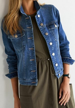 Femme portant une veste en jean bleue sur une robe vert olive avec une taille froncée, debout devant un fond clair uni.