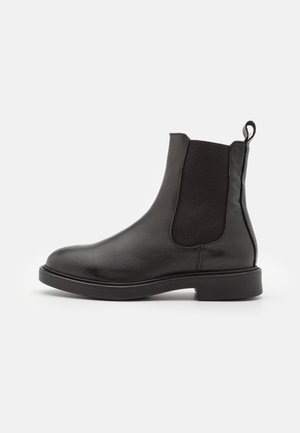 Bottine Chelsea en cuir noir avec des panneaux élastiques sur les côtés, bout arrondi et languette, présentant une texture lisse et une semelle plate en caoutchouc.