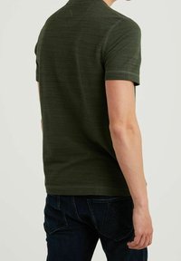 Camiseta corta de manga en verde oscuro con un patrón texturizado y corte ajustado, detalles de costura visibles, combinada con jeans de color azul oscuro.