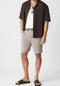 Hombre con camisa de manga corta marrón oscuro abotonada sobre camiseta blanca sin mangas, shorts beige con cinturón tejido, gafas de sol y sandalias oscuras tipo slide.