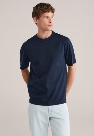 Jeune homme aux cheveux bouclés portant un t-shirt à manches courtes bleu marine et un jean bleu clair, regardant sur le côté avec les mains derrière le dos.