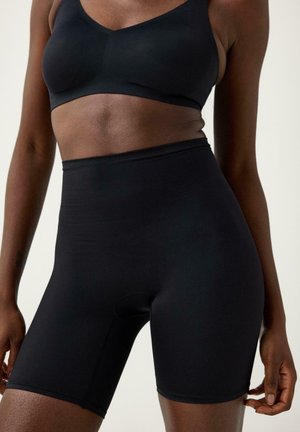 Vrouw die een zwart sportbeha en hoge zwarte compressiekorte broek draagt, gezien van het midden van de torso tot het midden van de dijen tegen een effen achtergrond.
