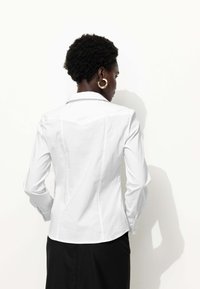 Chemise blanche ajustée avec un col pointu, des manches longues et des détails de couture. Tissu en coton lisse avec un design épuré et minimaliste.