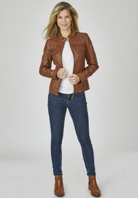 Braune Lederjacke mit Reißverschluss, Stehkragen und zwei Brusttaschen. Kombiniert mit blauen Jeans und braunen Stiefeletten.