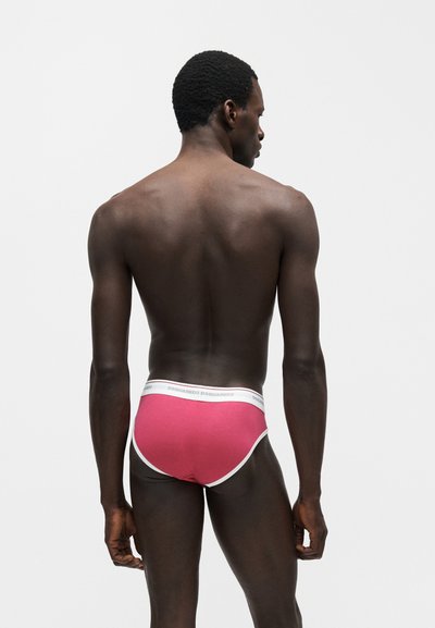Άνδρας με σκουρόχρωμο δέρμα γυρισμένος ανάποδα, φοράει κόκκινα εσώρουχα με λευκή ελαστική μέση και περιθώριο, στέκεται μπροστά σε λευκό φόντο.