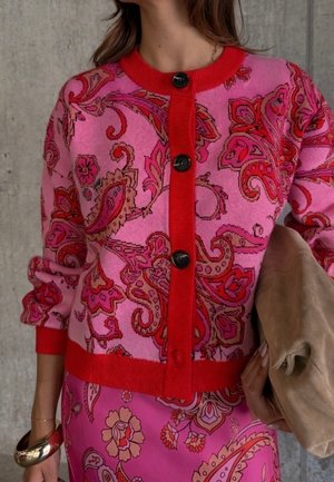 Kvinde iført en cardigan med pink og rød paisley-mønster og sorte knapper, matchende nederdel, guldbøjle og holder en tan clutch-taske.