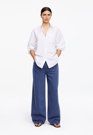 Kobieta stoi z rękami w kieszeniach, ubrana w luźną białą koszulę na guziki, szerokie niebieskie spodnie i czarne sandały na białym tle.