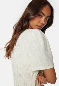 Abito bianco con maniche corte, decorato con dettagli in broderie anglaise, tessuto testurizzato e vita aderente. Superficie liscia e morbida con un sottile motivo.