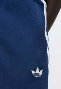 Tumši zilas kokvilnas sporta bikses ar baltām sānu svītrām un nelielu baltu Adidas logotipu, ar mīkstu tekstūru un redzamu kabatas detaļu.